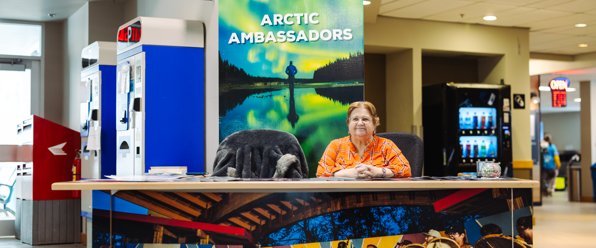 The image features a woman sitting at the arctic ambassador desk at yellowknife airport